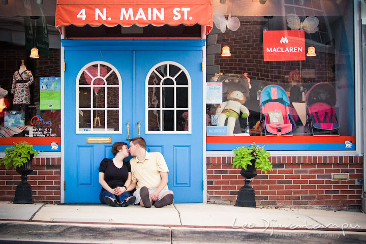 Engaged guy and girl sitting on the ground, kissing in front of a colorful store. Engagement Pre-wedding Photo Session Bel Air Maryland wedding photographer