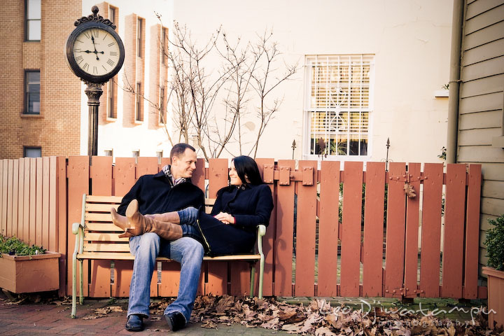 engaged couple sitting on bench, by a clock and orange fence gate. Urban City Pre-wedding Engagement Photographer Annapolis MD