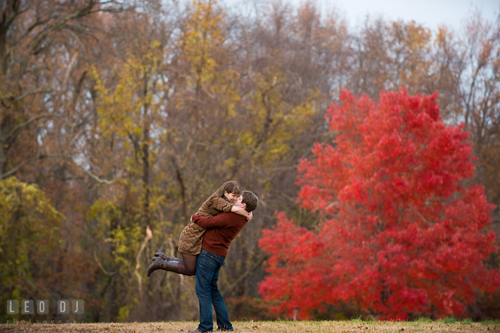 Engaged guy lifted up his fiancée on a field with red fall foliage trees in the background. Pre-wedding engagement photo session at Annapolis, Eastport, Maryland, Quiet Waters Park, by wedding photographers of Leo Dj Photography. http://leodjphoto.com