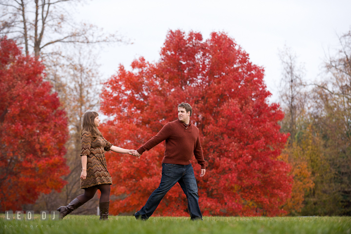Engaged couple walking together on a field with red fall foliage trees. Pre-wedding engagement photo session at Annapolis, Eastport, Maryland, Quiet Waters Park, by wedding photographers of Leo Dj Photography. http://leodjphoto.com