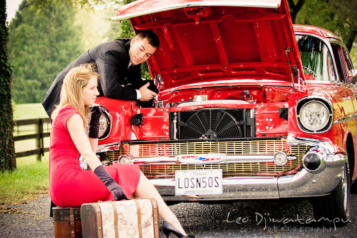 Engaged gentleman, opened front vehicle hood to check out engine while looking at his fiancee. Pre-wedding engagement photography session old antique Chevy Bel Air car, dress, outfit, accessories, suitcases
