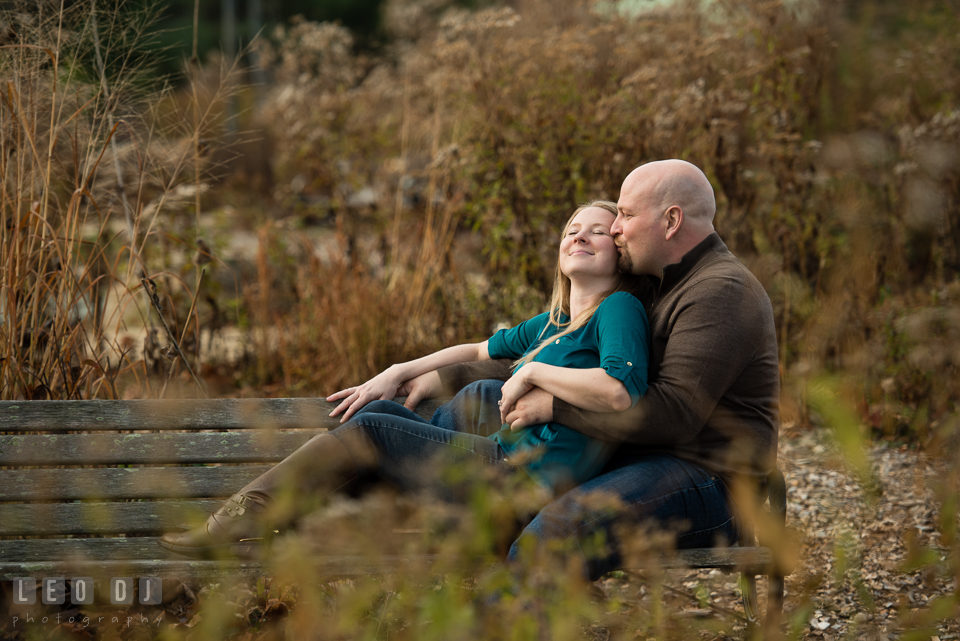 Quiet Waters Park Annapolis Maryland engaged man sitting on bench hugging and kissing fiancee photo by Leo Dj Photography.