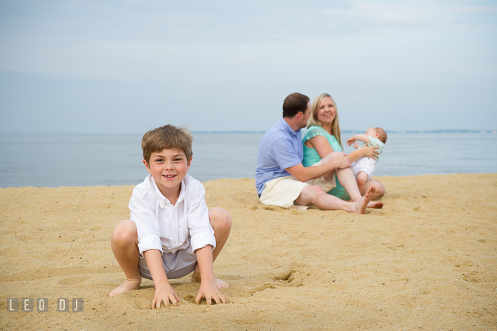 Boy smiling while Father, Mother and baby brother playing together in the background. Chesapeake Bay, Kent Island, Annapolis, Eastern Shore Maryland children and family lifestyle portrait photo session by photographers of Leo Dj Photography. http://leodjphoto.com