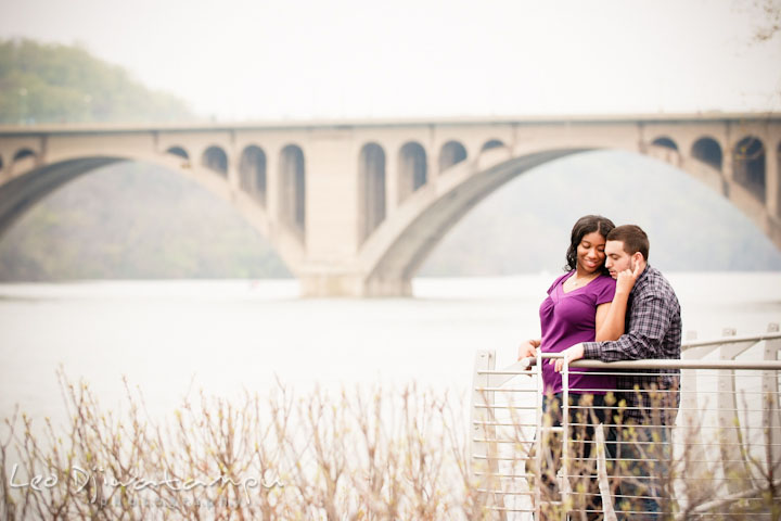 Engaged girl cuddled with her fiancé by the Francis Scott Key Bridge. Pre wedding engagement photo session at Georgetown, Washington DC by wedding photographer Leo Dj Photography