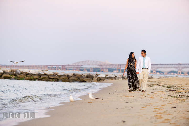 Engaged girl and her fiancé walking on the beach. Bay Bridge in the back ground. Indian pre-wedding or engagement photo session at Eastern Shore beach, Maryland, by wedding photographers of Leo Dj Photography.