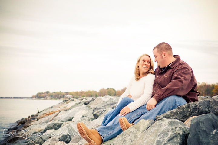 Fiancee couple smiling, sitting on rocks by water. Engagement Photographer Matapeake Beach, Chesapeake Bay