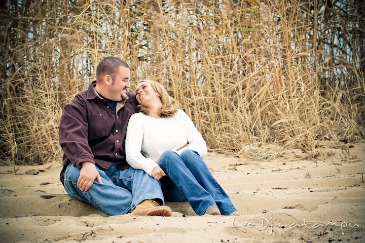 engaged couple cuddling, sitting on sand, grass straw background. Engagement Photographer Matapeake Beach, Chesapeake Bay