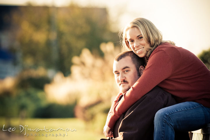 engaged girl sitting behind her fiancee guy, smiling, looking at camera. Engagement Photographer Matapeake Beach, Chesapeake Bay