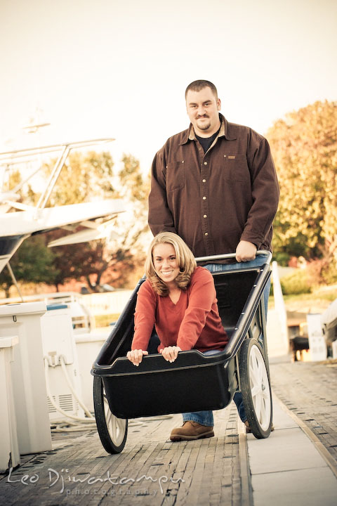 engaged couple posing. fiancee girl on cart. guy pushing cart on marina pier by Hemingways and Chesapeake Bay Beach Club. Engagement Photographer Matapeake Beach, Stevensville