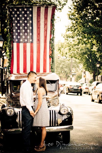fiancee couple holding hands kissing by antique car with American flag. fun candid engagement prewedding photo session Old Town Alexandria VA Washington DC
