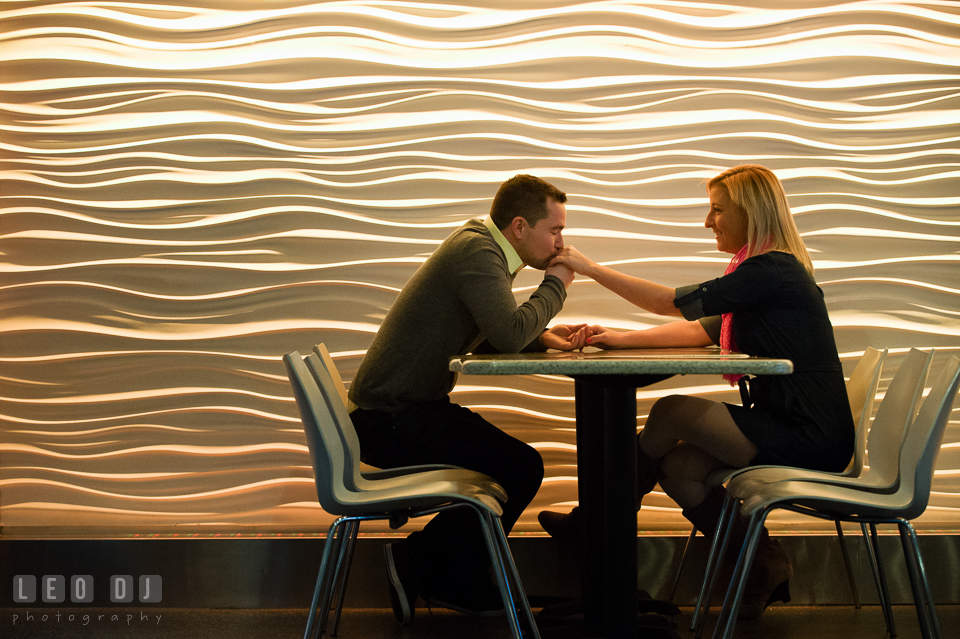 Engaged man siting by a table, kissing his fiancée's hand. Baltimore MD pre-wedding engagement photo session at National Aquarium in Baltimore, by wedding photographers of Leo Dj Photography. http://leodjphoto.com