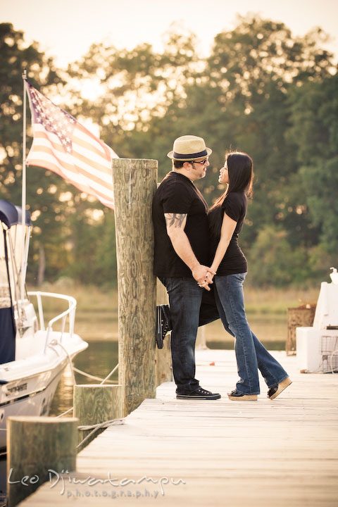 Engaged couple cuddling on the pier. American flag in the background. Eastern Shore MD engagement pre-wedding photo session pier boat tattoo