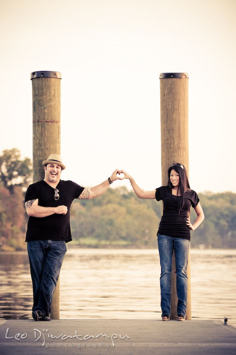 Engaged couple standing by the water, making heart symbol. Eastern Shore MD engagement pre-wedding photo session pier boat tattoo