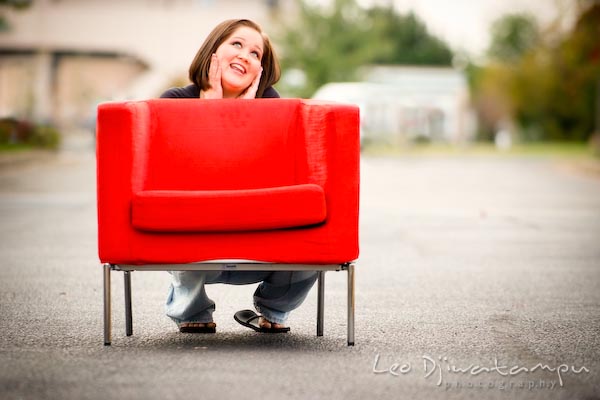 Girl squatting on ground behind red chair, posing, smiling. Commercial work photography Annapolis Eastern Shore MD Washington DC