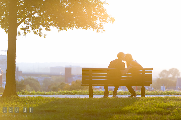 Engaged couple sitting on the bench beneath a tree, kissing. Engagement photo session at town home near Federal Hill Park Baltimore Maryland by wedding photographers of Leo Dj Photography (http://leodjphoto.com)