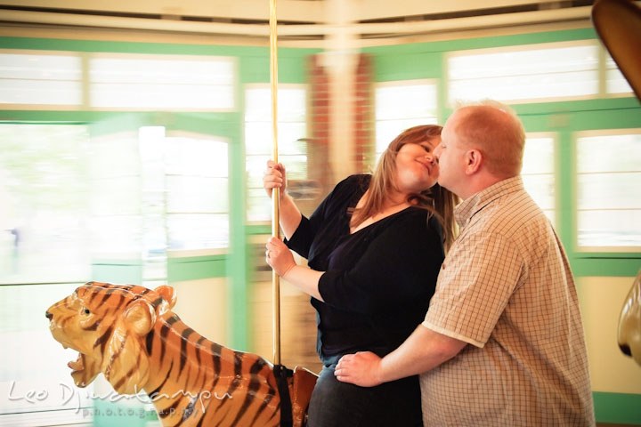 Engaged girl on carousel trying to kiss her fiancé. Pre wedding engagement photo session at Glen Echo Park Maryland by wedding photographer Leo Dj Photography