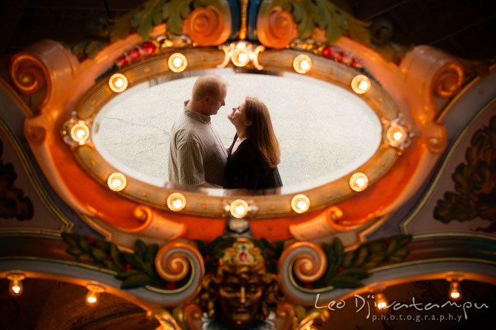 Engaged couple hugging, reflected on a carousel mirror. Pre wedding engagement photo session at Glen Echo Park Maryland by wedding photographer Leo Dj Photography