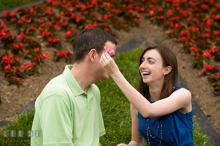 Engaged girl laughing trying to put flower on her fiancé's ear. Pre-wedding or engagement photo session at University of Maryland at College Park campus by wedding photographers of Leo Dj Photography.