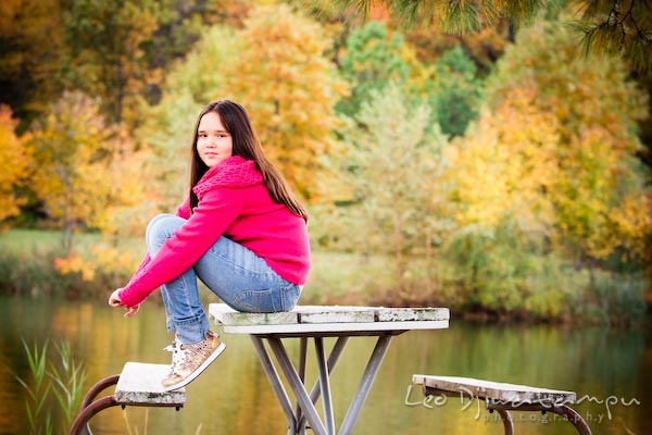 girl in pink sweater and jeans sitting on picnic table, by pond, fall colored trees. Commercial comp card photograper Kent Island Annapolis MD Washington DC