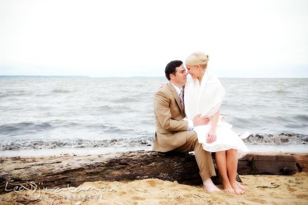 bride sit on groom's lap on a log at chesapeake bay beach, waves behind. Annapolis Kent Island MD Modern Intimate Candid Posed Photojournalistic Style Wedding Photographer