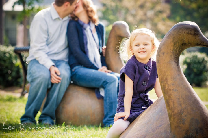 Little blonde girl looking at camera smiling while mom and dad kissing in the backround. Annapolis Maryland candid lifestyle family portrait photography