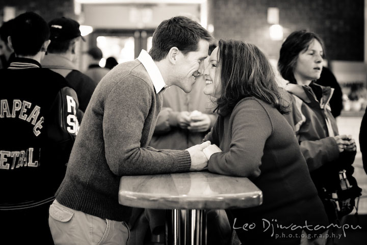 Engaged couple holding hands and smiling, people passing by at Market House. Urban city theme engagement session photographer Annapolis MD