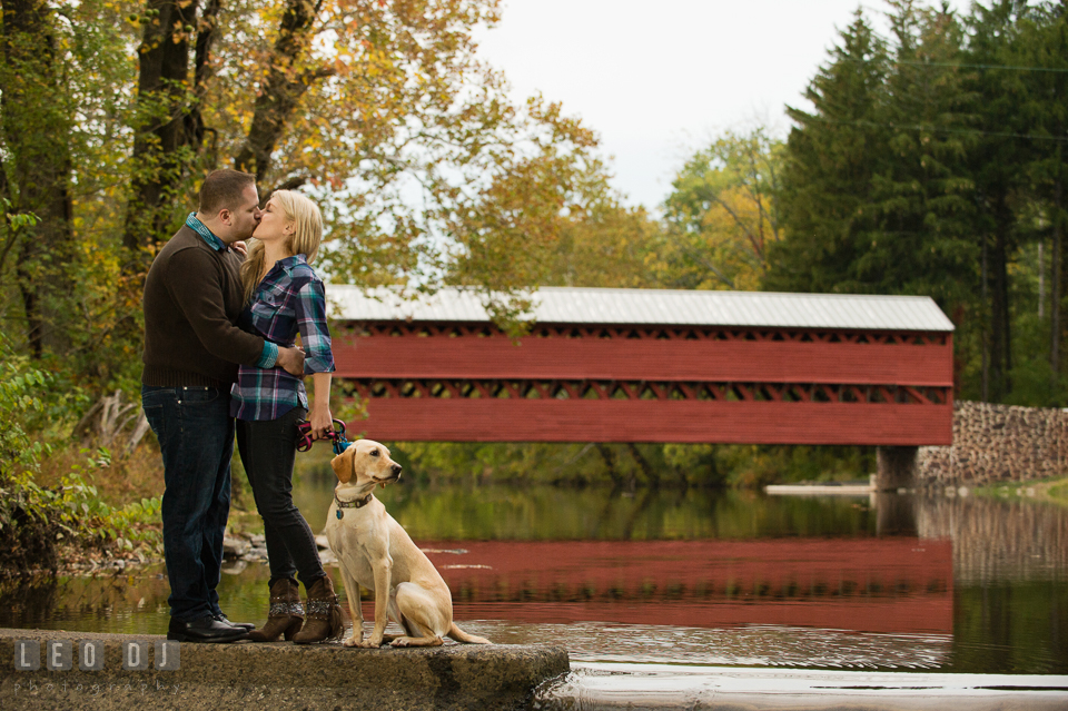 Engaged couple kissing by the river by the Sachs Covered Bridge with their labrador dog. Gettysburg PA pre-wedding engagement photo session, by wedding photographers of Leo Dj Photography. http://leodjphoto.com