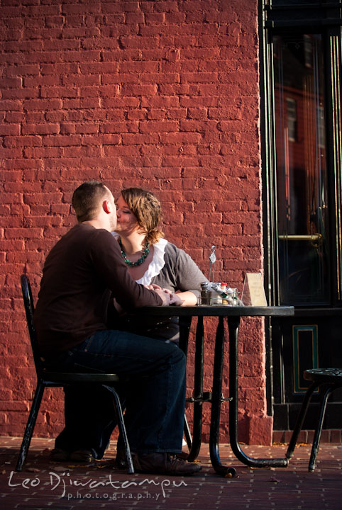 Engaged guy and girl sitting at an outside restaurant table and kissing. Pre-wedding or engagement photo session at Annapolis city harbor, Maryland, Eastern Shore, by wedding photographers of Leo Dj Photography.