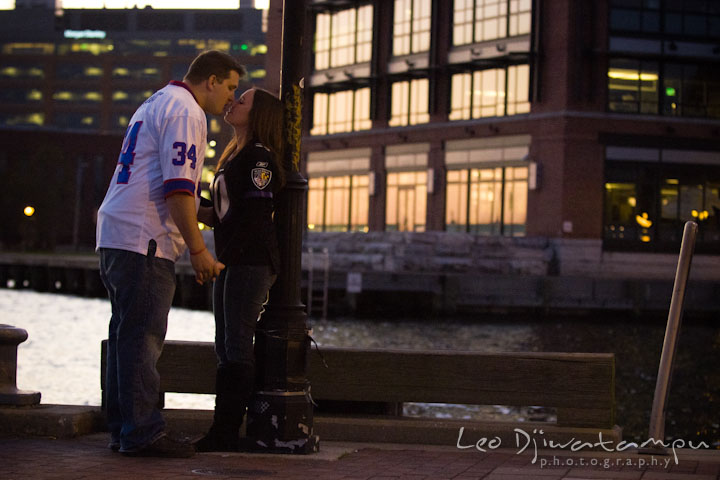 Engaged couple in Ravens football jerseys, kissing. Fells Point Baltimore Maryland pre-wedding engagement photo session with their dog pet by wedding photographers of Leo Dj Photography