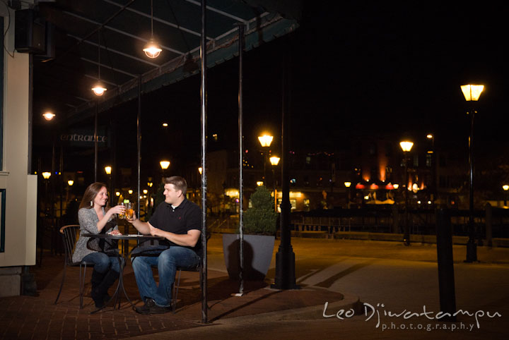 Engaged couple sitting outside a restaurant, raised their glasses of beers. Fells Point Baltimore Maryland pre-wedding engagement photo session with their dog pet by wedding photographers of Leo Dj Photography