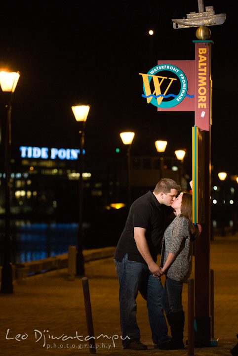 Engaged couple kissing by a Baltimore sign and Tide Point in the background. Fells Point Baltimore Maryland pre-wedding engagement photo session with their dog pet by wedding photographers of Leo Dj Photography
