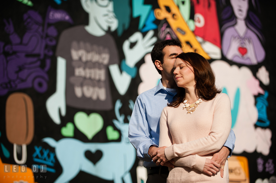 Engaged guy hugging his fiancé in front of a colorful wall mural. Washington DC pre-wedding engagement photo session at Adams Morgan and the Smithsonians, by wedding photographers of Leo Dj Photography. http://leodjphoto.com