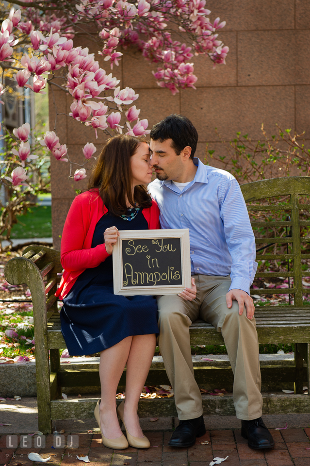 Engaged couple sitting on a bench under a magnolia tree holding a sign, See you in Annapolis for their weddingfiancé . Washington DC pre-wedding engagement photo session at Adams Morgan and the Smithsonians, by wedding photographers of Leo Dj Photography. http://leodjphoto.com