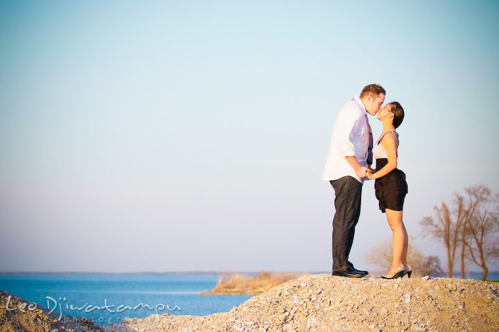 engaged couple holding hands and kissing, blue water in the background. Romantic engagement session on beach Kent Island Maryland