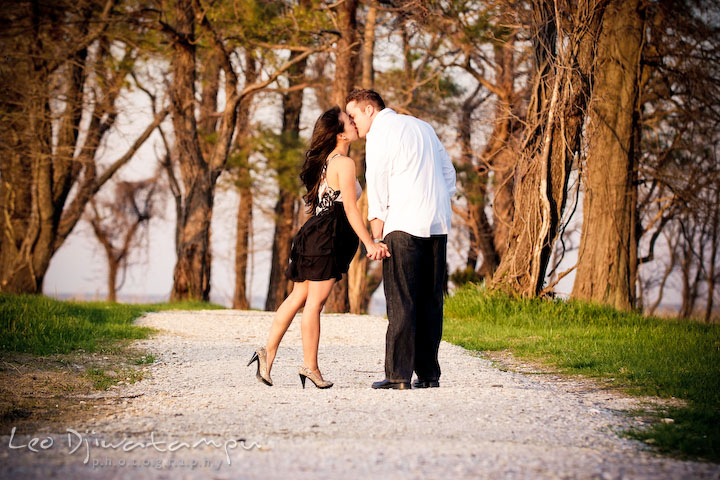 engaged couple walking down a gravel path in forest, holding hands and kissing. Romantic engagement session on beach Kent Island Maryland