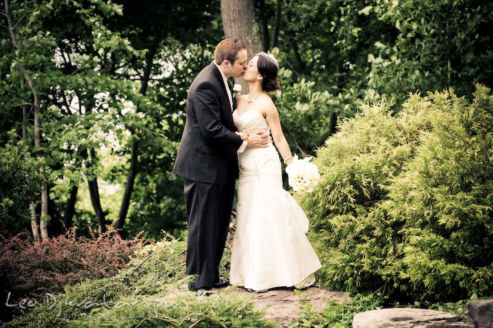 Bride and groom kissing by the green trees and bushes. Falls Church Virginia 2941 Restaurant Wedding Photography