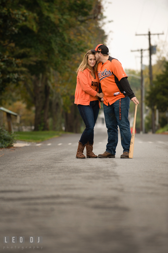 Engaged man, holding Louisville Slugger baseball bat, kissing his fiancee, both wearing Baltimore Orioles apparel. Eastern Shore Maryland pre-wedding engagement photo session at St Michaels MD, by wedding photographers of Leo Dj Photography. http://leodjphoto.com