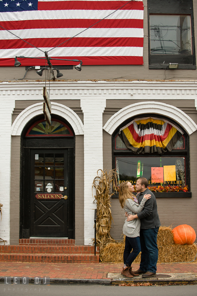 Engaged couple cuddling on the street with decors of pumpkin, bales of hay or straw, Maryland flag-color banner, and American flag. Eastern Shore Maryland pre-wedding engagement photo session at St Michaels MD, by wedding photographers of Leo Dj Photography. http://leodjphoto.com