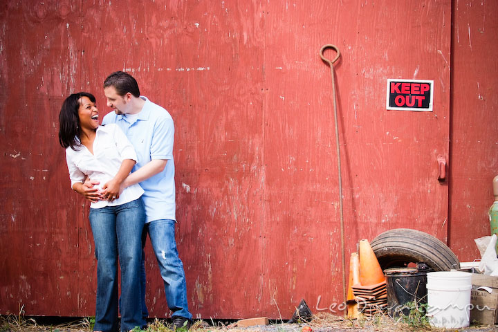 Engaged couple cuddling and laughing in front of a red barn wall with keep out sign. Engagement pre-wedding photo session fruit tree farm barn flower garden by Leo Dj Photography