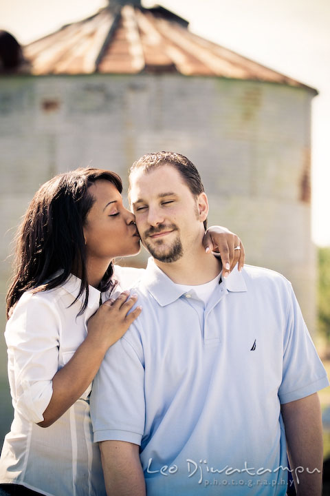 Engaged girl kissed fiancee's cheek. Grain silo behind them. Engagement pre-wedding photo session fruit tree farm barn flower garden by Leo Dj Photography