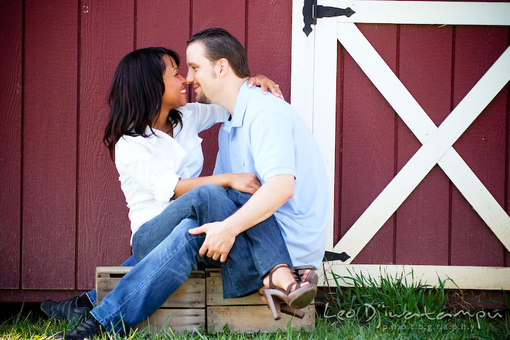 Engaged couple sitting close together and laughing by a red barn. Engagement pre-wedding photo session fruit tree farm barn flower garden by Leo Dj Photography