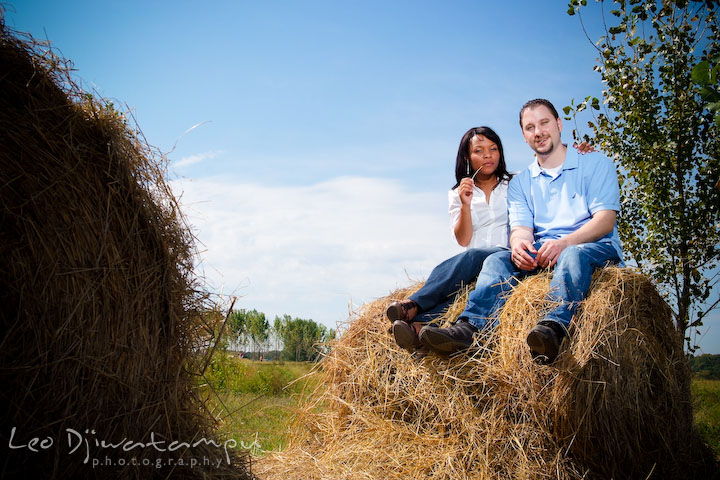 Engaged couple sitting on a hay roll. Engagement pre-wedding photo session fruit tree farm barn flower garden by Leo Dj Photography