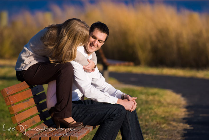Engaged guy sitting on the bench, hugged by his fiancee. Pre-wedding engagement photo session at Washington College and Chestertown, Maryland, by wedding photographer Leo Dj Photography.
