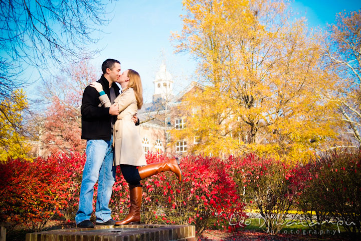 Engaged couple kissing in between yellow and red fall colored trees and bushes at campus. Chestertown Maryland and Washington College Pre-Wedding Engagement Session Photographer, Leo Dj Photography