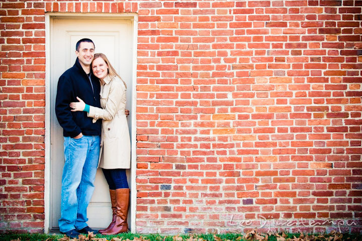 Engaged couple posing in front of a brick wall with a white door. Chestertown Maryland and Washington College Pre-Wedding Engagement Session Photographer, Leo Dj Photography