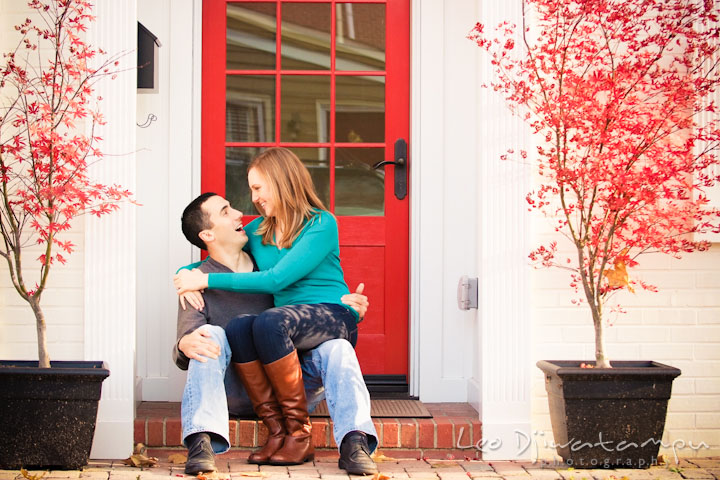 Engaged girl sitting on her fiancé's lap, in front of red door. Chestertown Maryland and Washington College Pre-Wedding Engagement Session Photographer, Leo Dj Photography