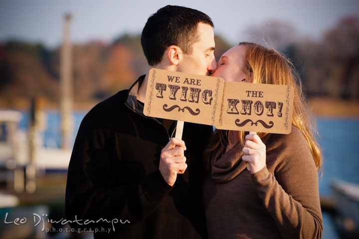 Fiancé and his fiancée kissing while holding we are tying the knot signs. Chestertown Maryland and Washington College Pre-Wedding Engagement Session Photographer, Leo Dj Photography