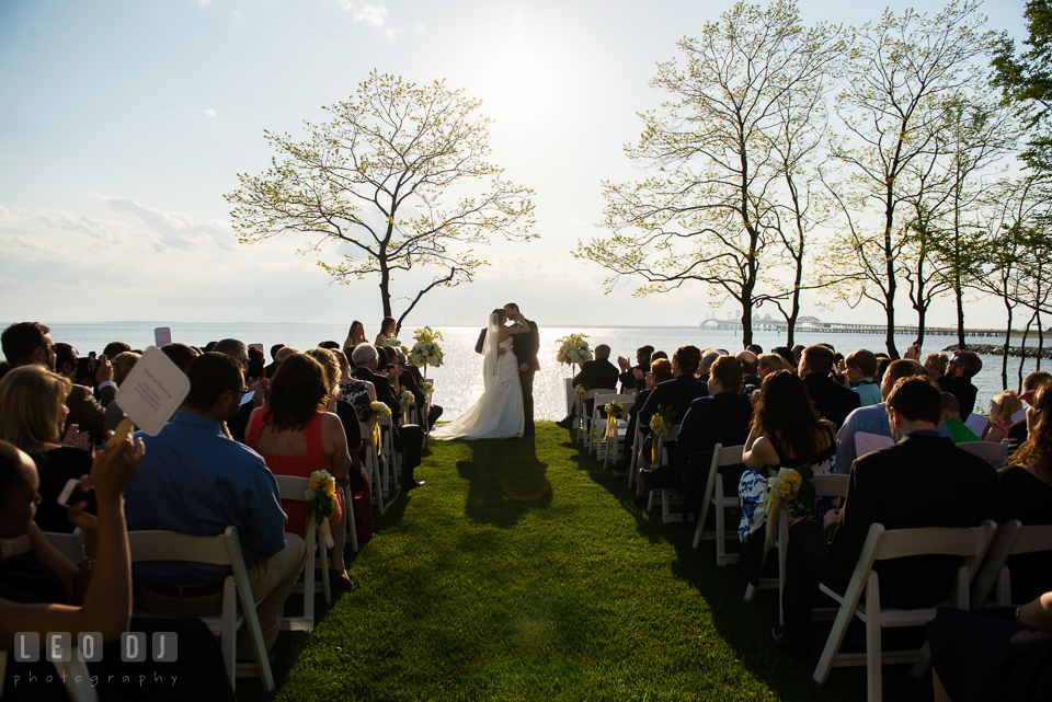 Bride and Groom kissing during the ceremony with the water and the Chesapeake Bay Bridge in the background. Kent Island Maryland Chesapeake Bay Beach Club wedding photo, by wedding photographers of Leo Dj Photography. http://leodjphoto.com