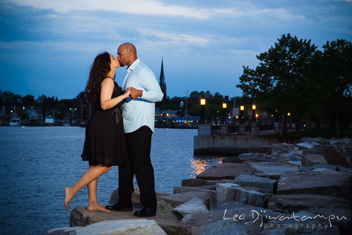 Engaged couple holding hand and kissing with Annapolis dowtown at night in the background. Annapolis Maryland USNA Pre-Wedding Engagement Photo Session by wedding photographer Leo Dj Photography