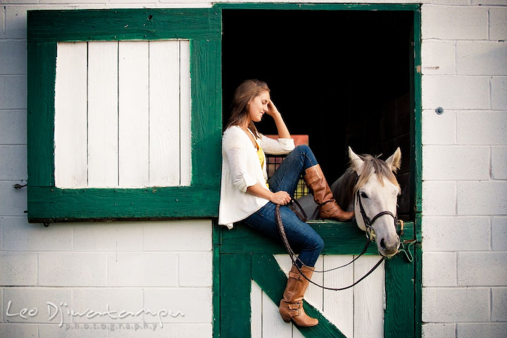 Girl sitting on stall door, brushing hair and smiling at her horse. Annapolis Kent Island Maryland High School Senior Portrait Photography with Horse Pet by photographer Leo Dj